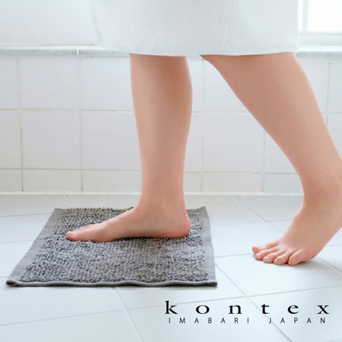 一個赤腳站在白色浴室地磚上的人，腳下踩著一塊灰色的 KONTEX 今治迷你快乾吸水浴墊，畫面呈現明亮簡約的衛浴空間感，並在右下角標註有 kontex IMABARI JAPAN 品牌字樣。