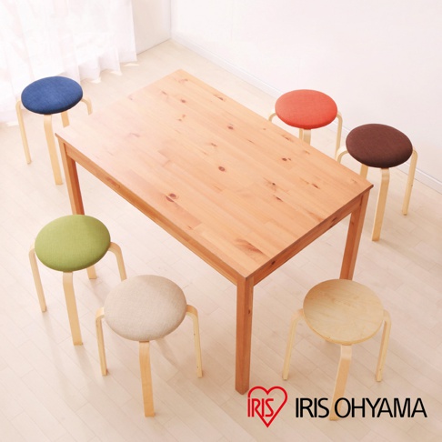 原木色木桌旁擺放著六張圓形木質椅凳，椅墊顏色包含藍色、綠色、米色、橘色、棕色及原木色，椅凳皆為三支木質椅腳。