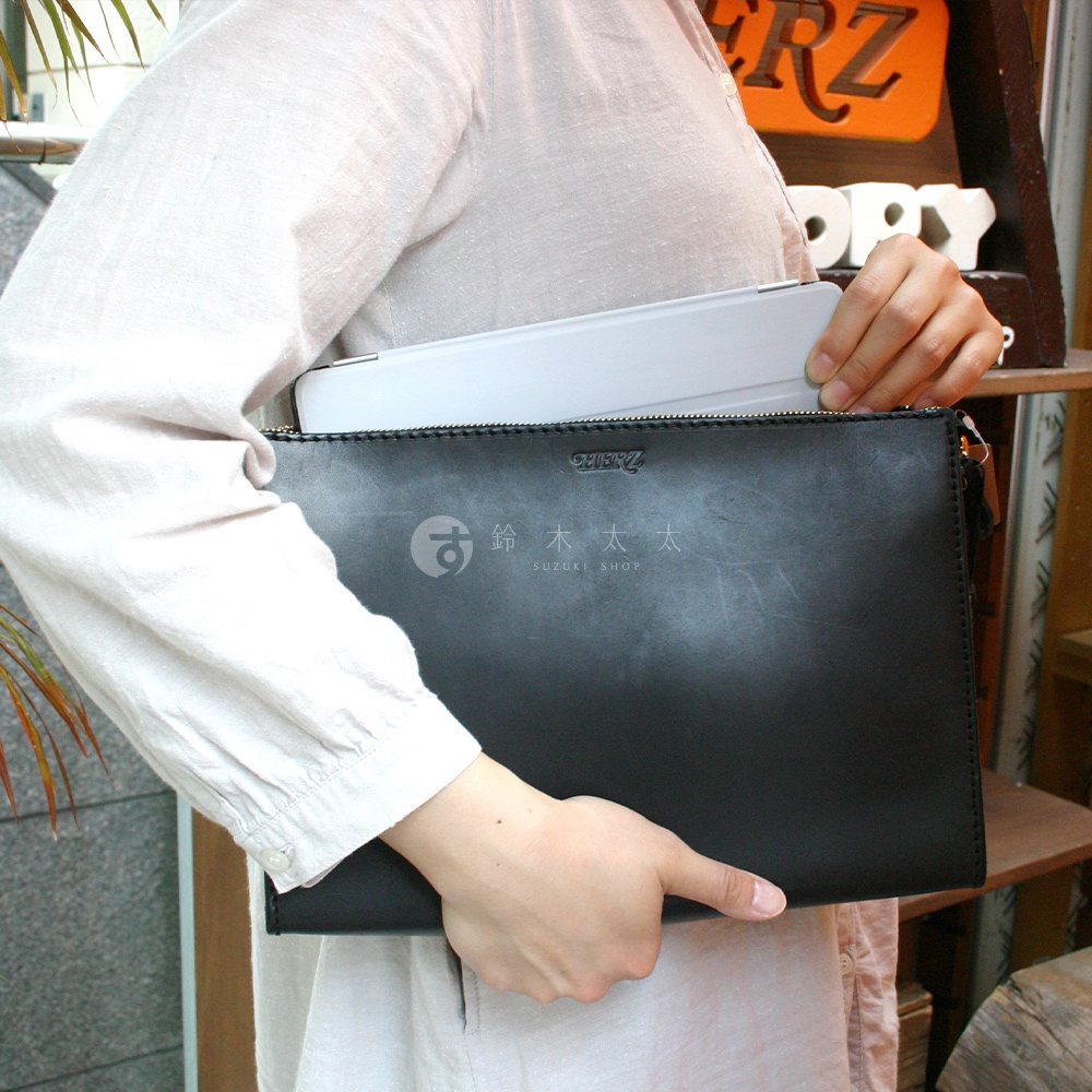 職人手作黑色皮革三角底拉鍊手拿包，包身印有「SUZUKI SHOP」品牌標誌，由一個人手持，包內可見一個白色平板電腦。