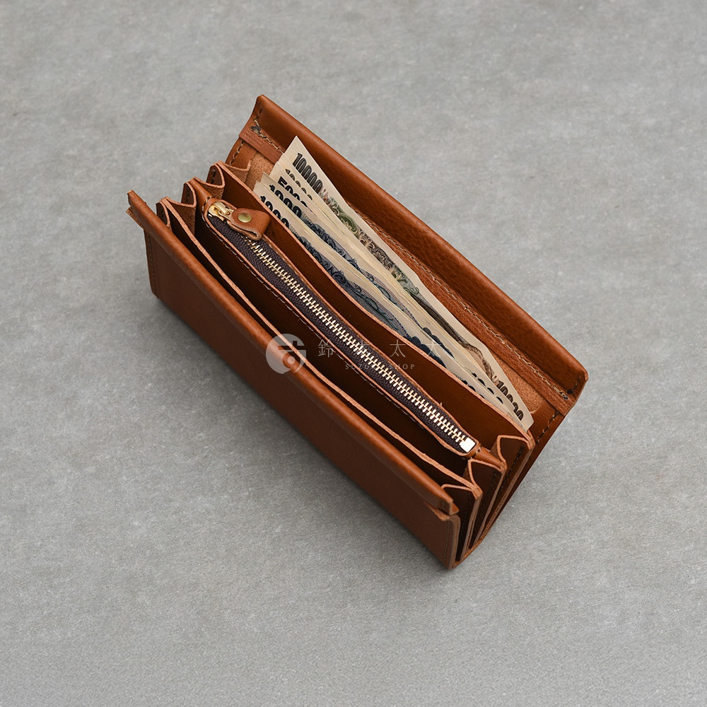 一款日本HERZ職人手作棕色皮革長夾，呈現半開狀態放置在灰色地面上，內部夾層裝有數張萬元與千元日幣紙鈔，並設有一個金屬拉鍊零錢袋，整體設計簡約具復古質感，中間隱約可見一個刻有品牌標誌的圓形圖案。