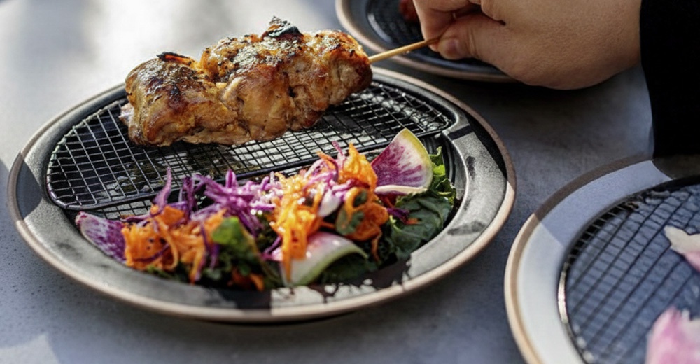 烤雞肉串放在烤網上，旁邊配有色彩繽紛的切絲沙拉，包含紫甘藍、胡蘿蔔和蘿蔔片。