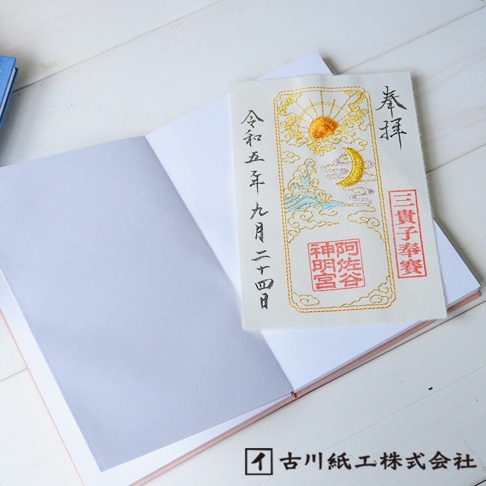 一本打開的御朱印帳，左頁為淺灰色空白頁，右頁上方疊放著一本白色御朱印貼紙，貼紙上有金色刺繡的太陽、彎月、波浪和雲朵圖案，並有日文文字與紅色印章。