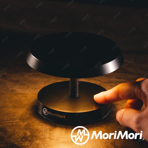 一個黑色圓形底座的 LED 造型燈，頂部有一個較大的黑色圓形燈罩，中間由一個銀色金屬桿連接。底座上印有 MoriMori 品牌標誌。一根手指正輕觸底座。