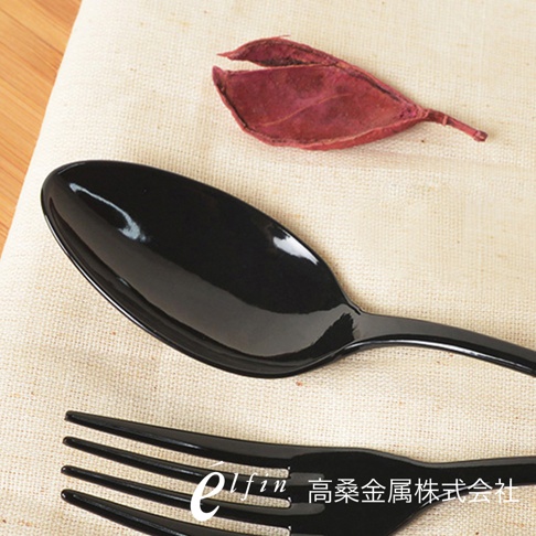 兩把日製黑色琺瑯餐匙，勺面光滑，呈現光澤感，置於米白色麻布上，旁邊點綴著一片乾燥的深紅色葉子。