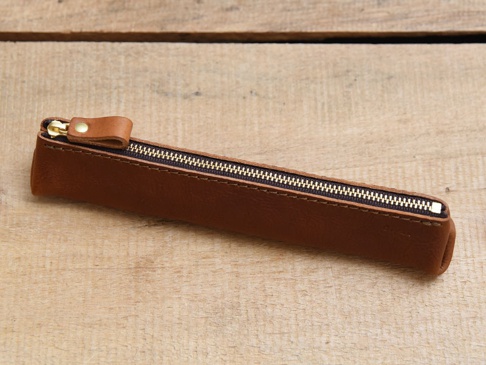 深棕色皮革製成的三角收納筆袋，配有金屬拉鍊和皮革拉袢，置於木紋背景上。