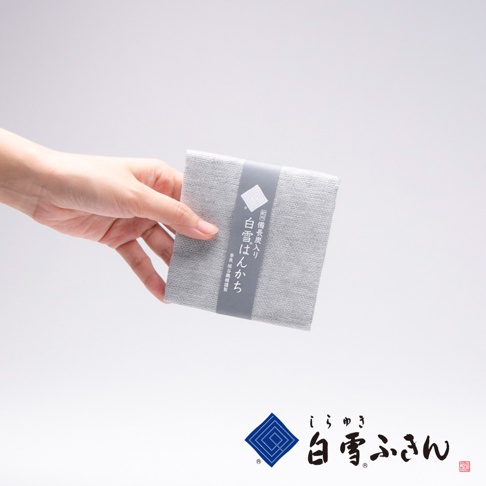 一隻手拿著一塊淺灰色、帶有細緻紋理的紀州備長炭方巾，方巾被一條深灰色紙帶包裹，紙帶上印有白色圖案和日文文字。
