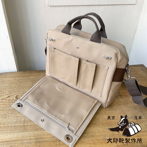 米色純棉帆布製成的橫式側背包，配有深棕色提把、肩帶及側邊皮飾。包身外側有四個淺色小口袋，內側翻蓋處有一個拉鍊口袋。