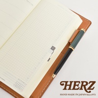 職人手作皮革旅行記事本書套，淺棕色皮革，內含筆記本，筆套中插著一支綠色與黑色筆身的鋼筆。