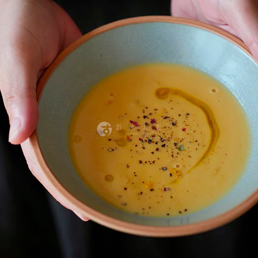 雙手捧著一個淺綠色內裡、陶土色外緣的陶瓷碗，碗中盛有濃稠的黃色湯品，表面撒有黑胡椒粒、紅色香料粒及淋上橄欖油。