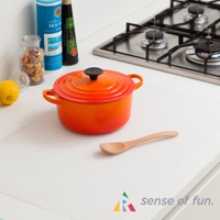 白色防滑料理台保護墊，墊上擺放著一個橘紅色圓形鑄鐵鍋，鍋蓋中央有黑色提把，鍋旁有一支木製湯匙，背景為廚房爐灶與調味品。