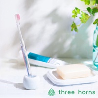 透明柄前傾式尖頭牙刷，帶粉紅色刷毛，立於白色牙刷架中，背景有牙膏、肥皂及綠葉裝飾。