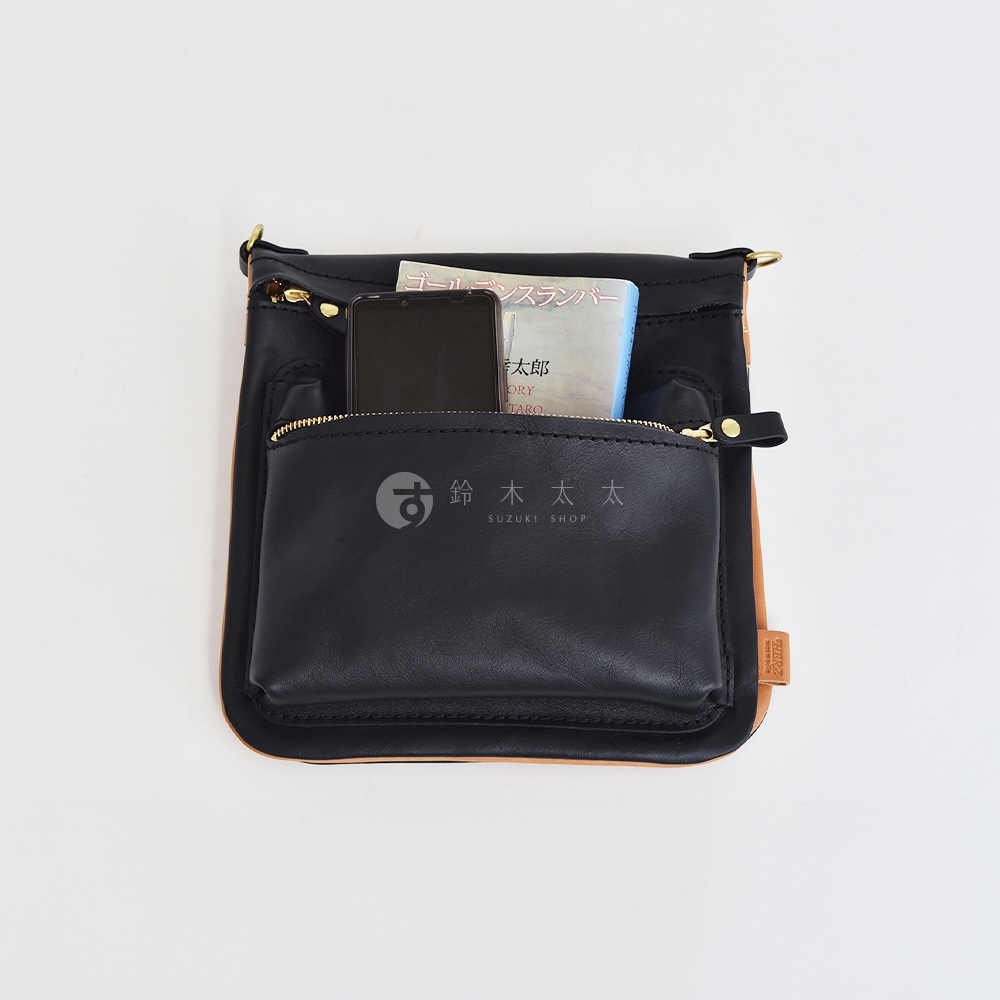 黑色皮革翻轉雙色直式斜背包，外部有拉鍊口袋，內部可見手機及書籍，配有金色五金和皮革掛繩。