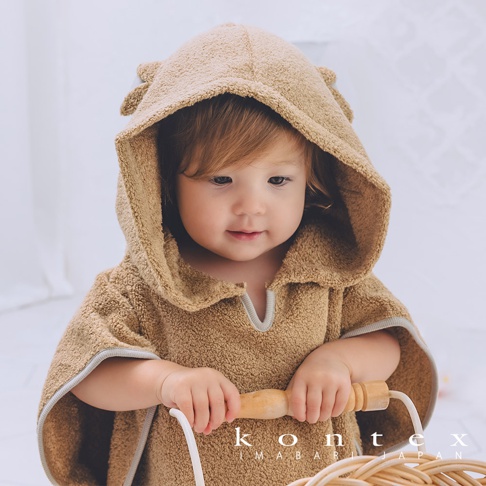 幼童身穿一件棕色絨毛材質的斗篷式連帽浴巾，帽子上有可愛的熊耳朵造型，浴巾邊緣有淺灰色滾邊。