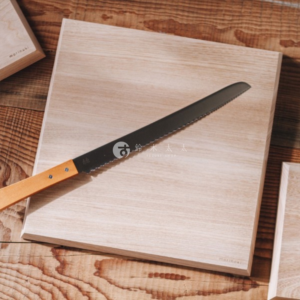 志津匠 morinoki 榆木砧板 S 尺寸，搭配一把黑色刀刃的麵包刀，砧板和木質桌面呈現自然紋理