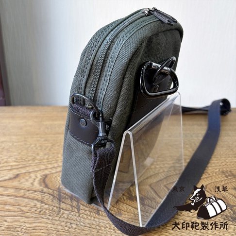 這款犬印鞄製作所的黑犬印石洗帆布斜背手機包，呈現軍綠色調，採用耐用的帆布材質搭配黑色皮革飾片與金屬五金配件。側面配有可拆卸肩背帶及黑色登山扣環，底部設有雙層拉鍊開口設計。包身由壓克力架支撐立於木紋桌面上，右下方印有品牌標誌與東京淺草犬印鞄製作所字樣。