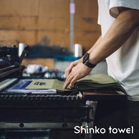 一疊軍綠色素面毛巾，由一位身穿白色T恤、手戴黑色智慧手錶的人在機器旁整理。