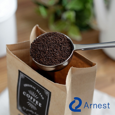 金屬咖啡量匙裝滿深棕色咖啡粉，置於一個打開的牛皮紙咖啡袋上方。