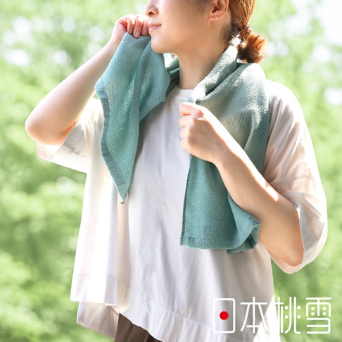 一位女性正將一條淺綠色的日本桃雪 NUGUW 今治輕量快乾運動長毛巾掛在頸部，她穿著白色寬鬆短袖上衣，背景為明亮的戶外綠意，圖片右下角有日本桃雪的品牌標誌。