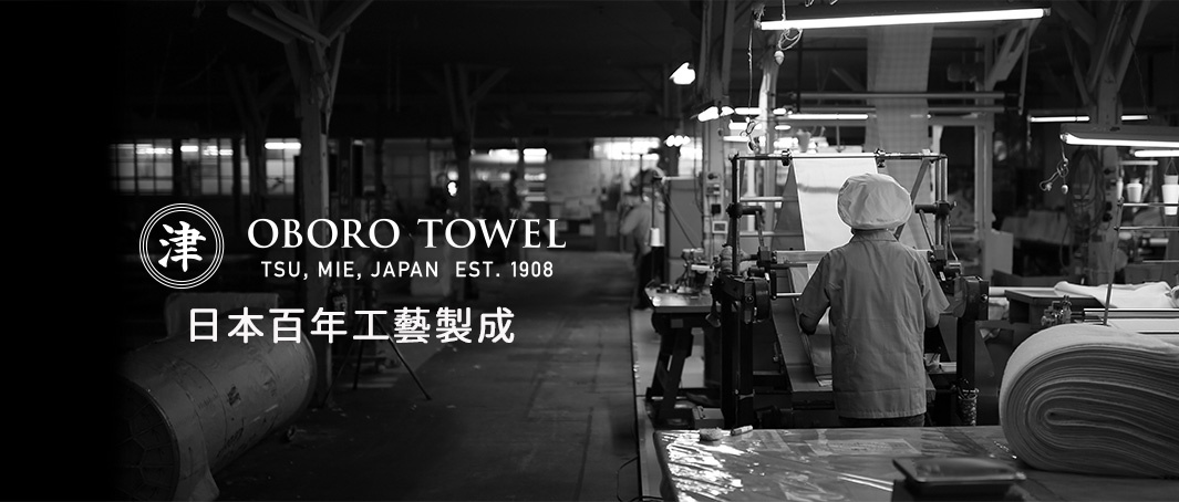 黑白攝影視角的日本 Oboro Towel 毛巾工廠內部，一名戴著白色工帽的工作人員正專注操作機台，背景整齊陳列著生產設備與紡織捲筒，畫面中央顯示 Oboro Towel 津、三重、日本及成立於 1908 年的品牌標誌與日本百年工藝製成字樣。