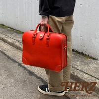 手提紅色皮革公事包，有金色旋釦和拉鍊，提把和側邊有金屬環，包包呈現方形，角落有縫線裝飾。