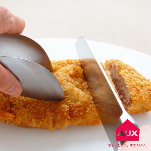 銀色金屬不沾桌調理幫手夾，夾著一塊炸至金黃的炸豬排，刀子正切斷豬排。