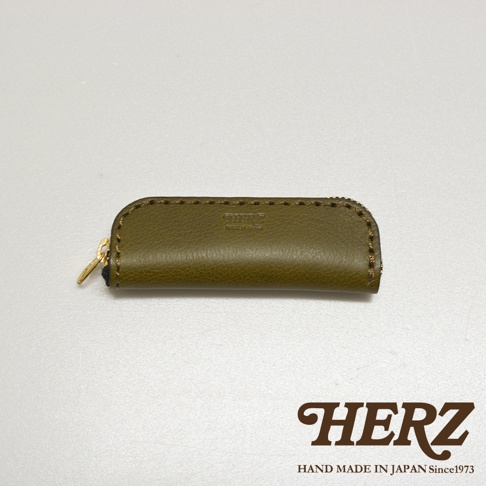 橄欖綠皮革製成的職人手作印章收納袋，配有金色拉鍊，袋身壓印有品牌標誌。