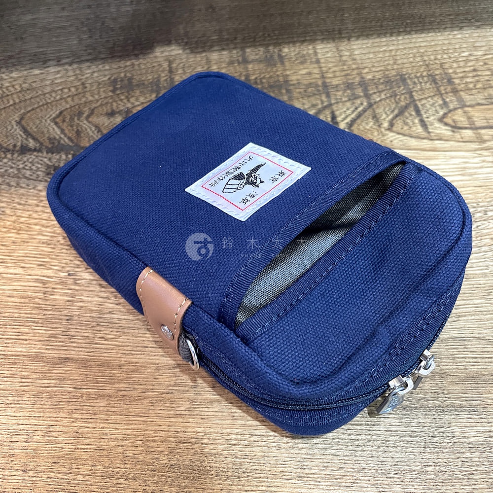 深藍色石洗帆布斜背手機包，有淺棕色皮革飾邊，正面有拉鍊開口和一個小口袋，上面繡有白色標籤。