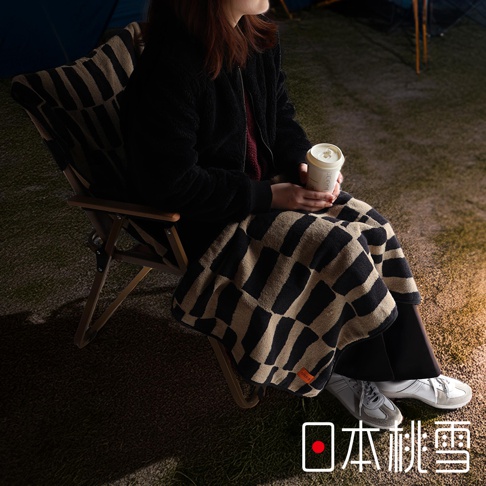 一位女子坐在戶外椅子上，蓋著日本桃雪的格紋毛毯，手裡拿著咖啡杯。