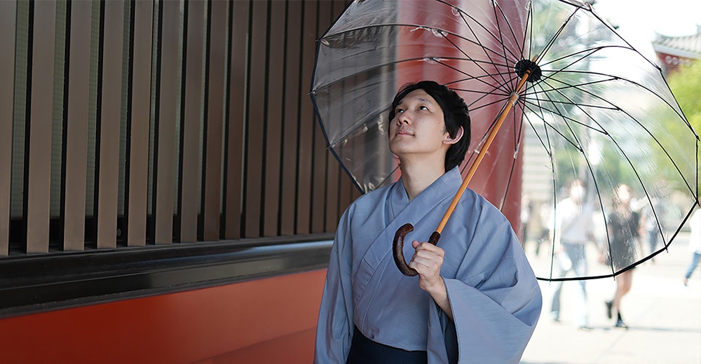 一位男子身著淺藍色和服，手持職人高級樺木透明傘，傘面由多根黑色傘骨支撐，傘柄為彎曲的樺木材質。