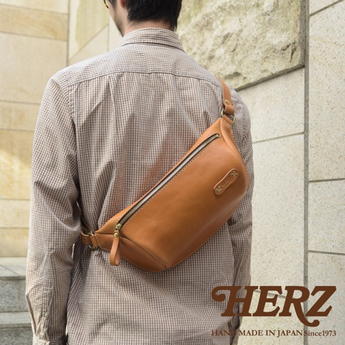 職人手作淺棕色皮革輕便斜背包，側面為圓潤弧形，頂部配有黃銅色拉鍊，右側有品牌皮標，側邊附有可調整的皮革揹帶。