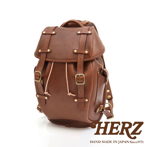 職人手作皮革經典雙扣後背包，呈現棕色澤，採用真皮材質，設有雙扣設計、抽繩束口及多個金屬扣環，風格復古。