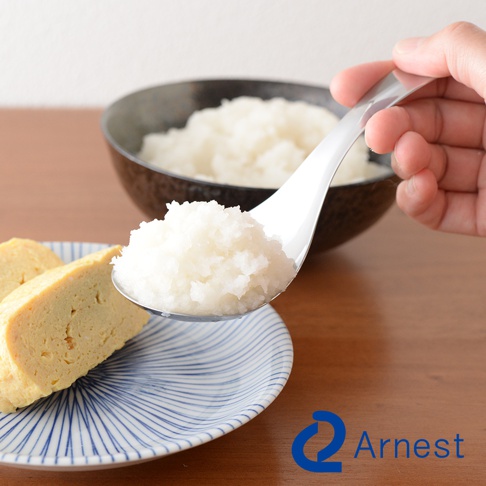 逸品物創不鏽鋼瀝水湯匙，舀起白色碎狀食材，湯匙背面有品牌 Logo，背景為盛有米飯的碗及盤中的玉子燒。