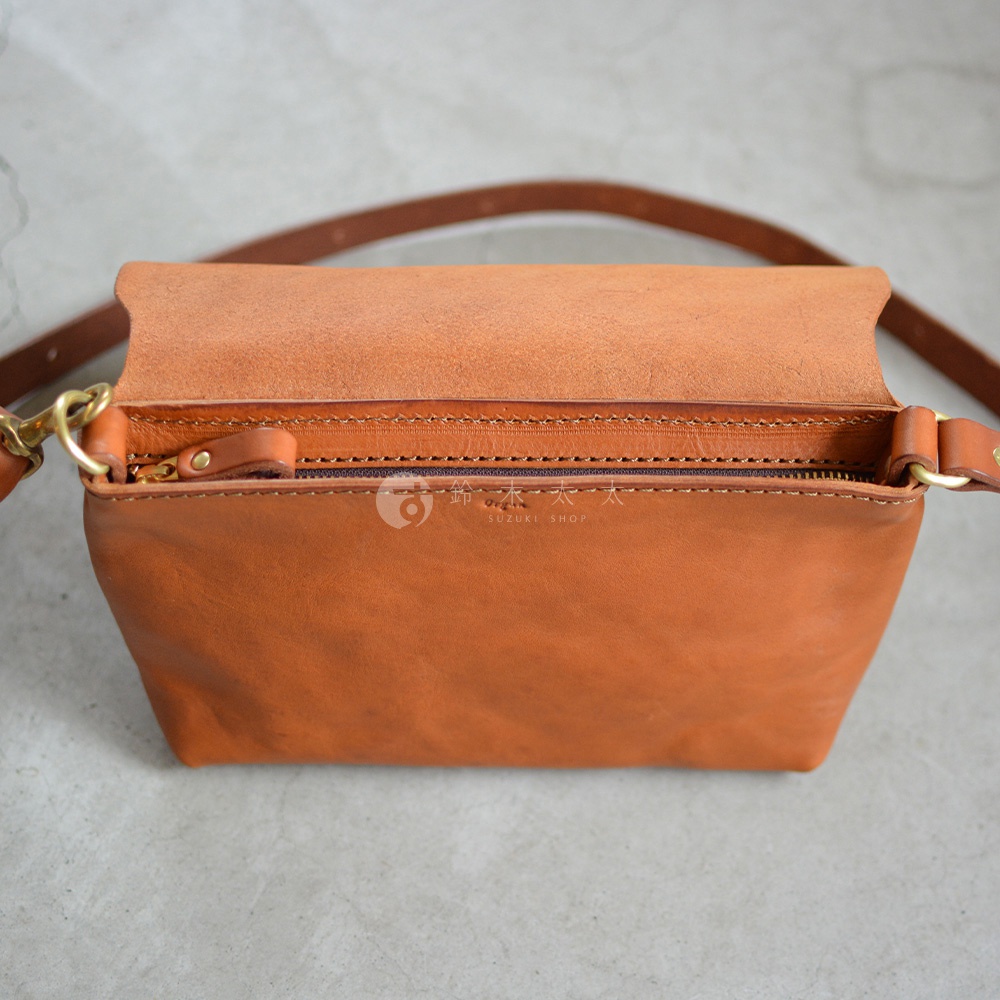 職人手作皮革隨身斜背包，採用棕色皮革製成，包身方正，頂部翻蓋設計，露出拉鍊開口，搭配細長皮質揹帶。