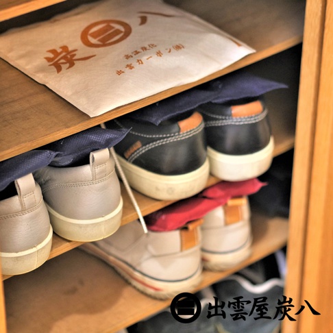 木質鞋櫃層架上整齊擺放著多雙休閒鞋，鞋內及上方分別放置了印有出雲屋炭八品牌標誌的除濕炭包，包含米白色、深藍色與紅色的不織布包裝，展現居家空間防潮除臭的生活應用場景。