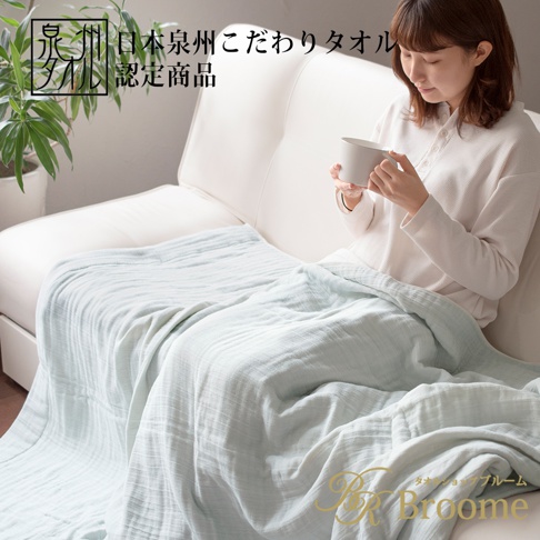 一位女性坐在白色沙發上，身上蓋著一條淺藍色、輕盈的四層紗毛巾毯。毛巾毯呈現柔軟皺褶的紋理。