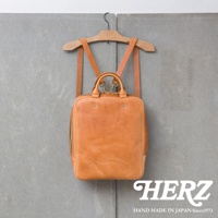 一個 tan 色皮革後背包，掛在木製衣架上，背包上有提把和可拆卸的肩帶，背景是灰色的牆壁，右下角有 HERZ 的標誌。