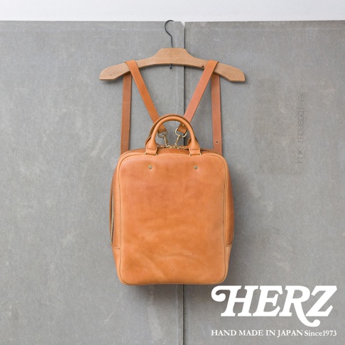 一個 tan 色皮革後背包，掛在木製衣架上，背包上有提把和可拆卸的肩帶，背景是灰色的牆壁，右下角有 HERZ 的標誌。