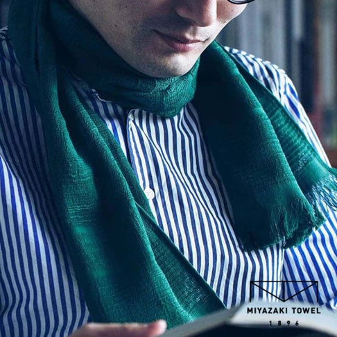 一位男士頸部圍繞著一條深綠色、帶有流蘇的今治匹馬棉圍巾，圍巾材質輕薄，露出內搭的藍白條紋襯衫。