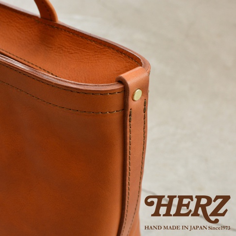 HERZ 品牌標誌，下方有 HAND MADE IN JAPAN Since1973 字樣，圖片右側為手提包的皮革邊緣細節，以特寫方式呈現。