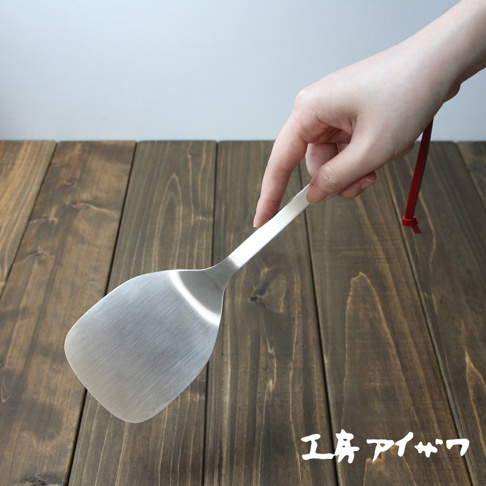 一只手持銀色不鏽鋼萬用鍋鏟，鍋鏟為扁平寬口設計，手柄細長，末端有紅色掛繩。鍋鏟置於深棕色木紋桌面上。