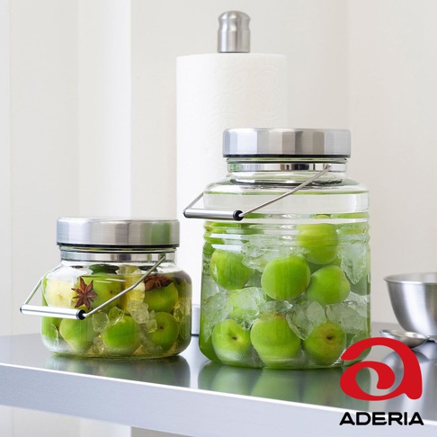 兩個透明玻璃梅酒罐，均配有不鏽鋼蓋和提把。左側罐子較小，內含青梅、冰塊、八角和薑片。右側罐子較大，內含青梅和冰塊，部分罐內液體呈現淡綠色。背景為白色，有捲筒紙巾和金屬碗。