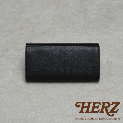 一張置於灰色素色背景上的黑色HERZ職人手作皮革長夾，呈現正面視角，皮夾材質為荔枝紋理皮革，表面質感細緻，下方印有棕色的HERZ品牌標誌以及HAND MADE IN JAPAN Since1973字樣。