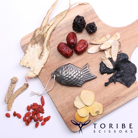 木製砧板上擺放著金屬製的魚形補鐵器，周圍散佈著乾燥的紅棗、枸杞、黑棗、人參鬚、當歸片和黑色的藥材。