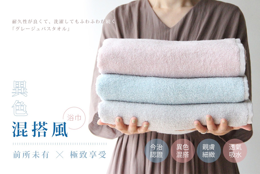 一位女士雙手疊放三條柔棉浴巾，顏色由上至下分別為淺粉色、淺藍色和淺灰色。浴巾材質呈現蓬鬆柔軟的織紋。