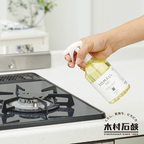 手持一瓶 SOMALI 泡沫式廚房清潔劑，瓶身為透明塑膠，內含淡黃色液體，白色噴頭。清潔劑瓶身貼有白色標籤，印有品牌名稱、產品名稱「KITCHEN CLEANER」及日文說明。清潔劑瓶口正對著黑色玻璃爐台。