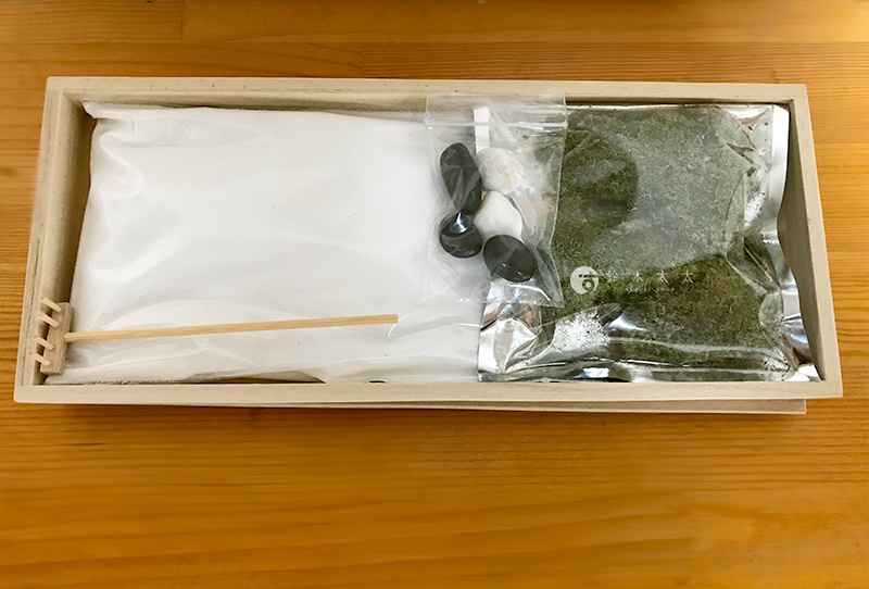 長方形木製托盤內裝有 zen garden 青苔庭園模型材料，包含一大包白色細沙、一包綠色人造青苔粉末、一小袋黑白相間的圓滑鵝卵石，以及一支精緻的迷你木製耙子。