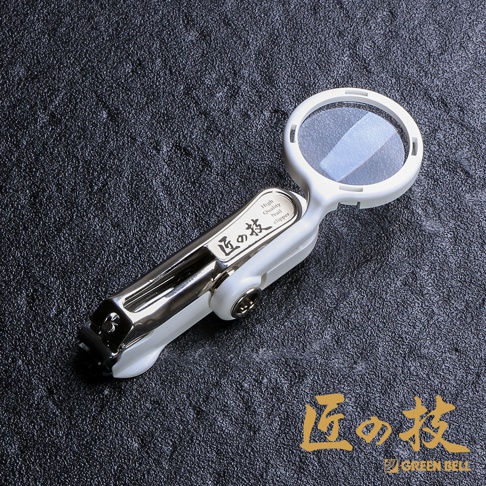 白色不鏽鋼附放大鏡指甲剪，放大鏡部分為圓形，帶有透明鏡片，指甲剪主體為銀色不鏽鋼與白色塑料組合，上面印有日文「匠の技」及英文「High Quality Nail clipper」字樣。