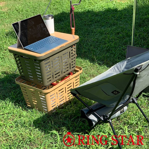 綠色和卡其色塑膠籃堆疊，上方綠色籃子上有木紋桌板，擺放著筆記型電腦和保溫杯，旁邊有一張綠色摺疊椅，背景為草地。
