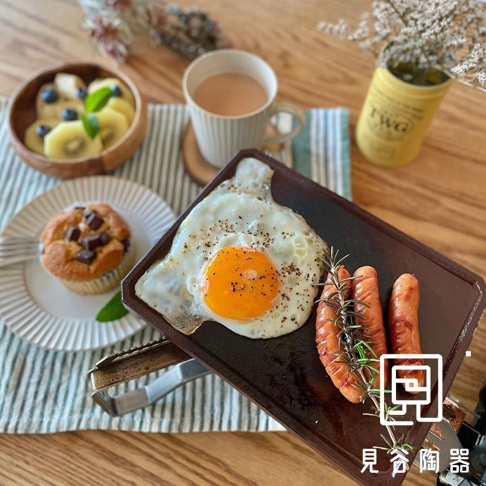 長方形的深棕色烤盤上，擺放著煎蛋、兩根香腸和迷迭香。烤盤旁邊有香濃咖啡、巧克力瑪芬與水果杯。