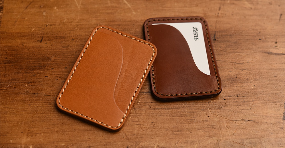 兩個職人手作皮革名片夾，一個為淺棕色，另一個為深棕色，皆縫有白色縫線，放在木質桌面。